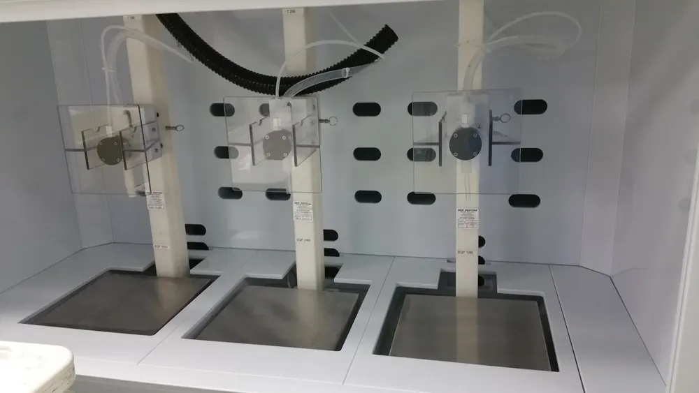 Three matched dispensing stations inside a white containment booth, each with a clear polycarbonate guard, feed tubing and a flush-mounted stainless platform recessed into the bench.