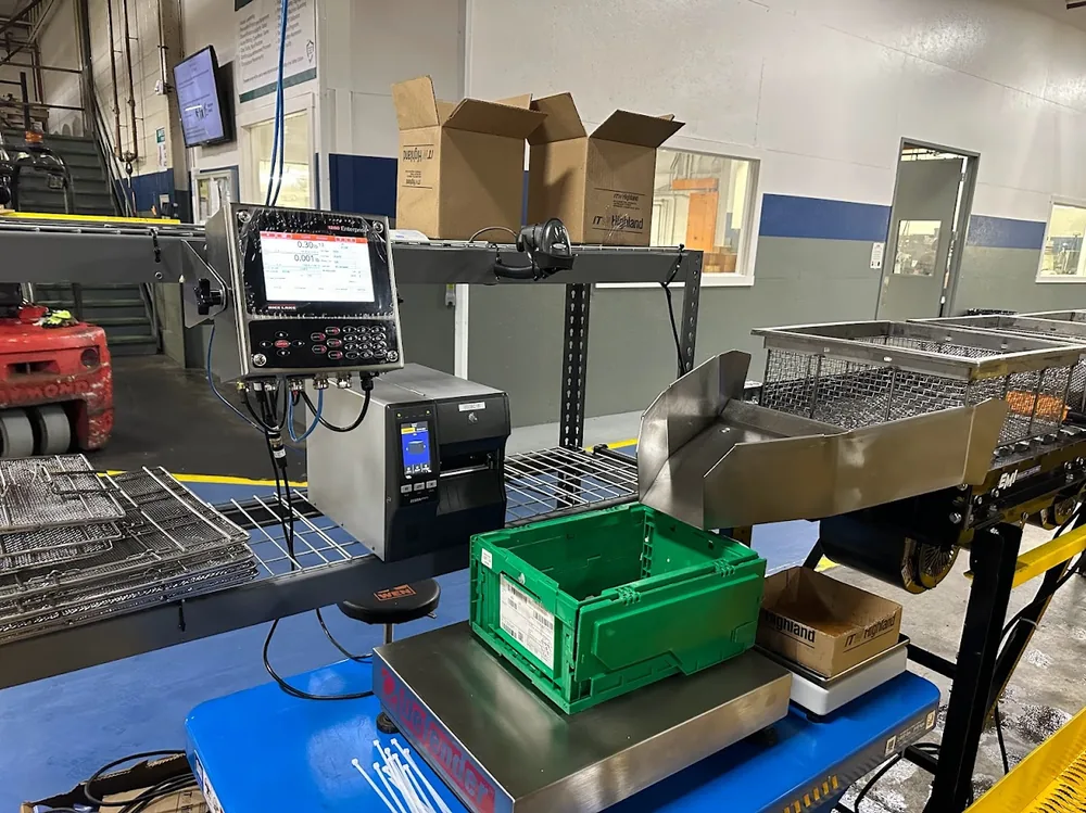 A scale indicator and Zebra industrial printer mounted over a floor scale weighing a green tote, with a barcode scanner alongside.
