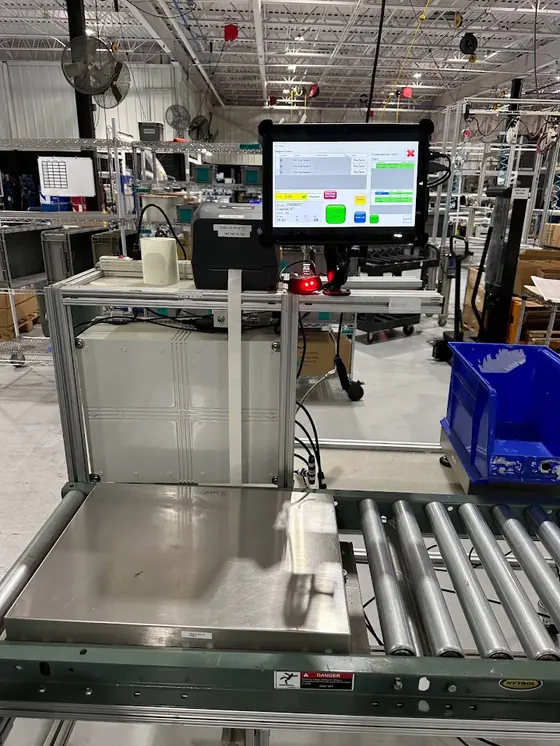 An in-line weighing station built into a roller conveyor: a stainless weigh platform flush with the rollers, a rugged touchscreen terminal, a Zebra label printer and a barcode scanner on an aluminium frame, with the control enclosure mounted beneath.