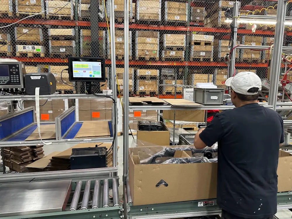 An operator boxing parts at a conveyor-side packing station, with a touchscreen terminal, scale indicator and Zebra label printer mounted at the line.