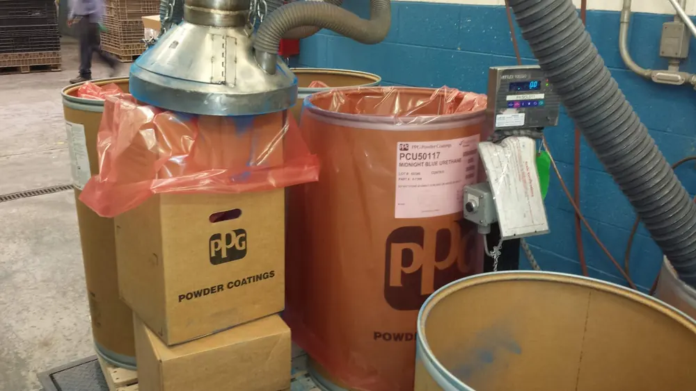 A powder coating drum being filled by weight under a stainless vacuum transfer hood, with a digital scale indicator reading zero mounted at the station.
